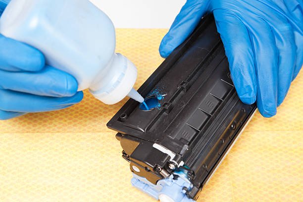 Gloved hands are adding cyan toner to a laser printer cartridge which is being refilled. Toner replacement is environmentally friendly, reducing cartridge waste. Toner refilling is also more cost effective and can be done by the end user with minimal equipment and skill.
